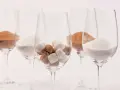 Cinq verres à vin contiennent différentes formes de sucre : du sucre en poudre blanc, du sucre brun en poudre et des morceaux de sucre blanc et brun. Une métaphore visuelle du sucre dans le vin.
