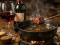Un morceau de bœuf est plongé dans une fondue fumante, accompagné d'un verre de vin rouge.
