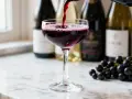 Un verre de vin rouge est servi, avec des grappes de raisin noir à côté. Le vin aligoté, souvent blanc, ici rouge, évoque la fraîcheur et la vivacité.