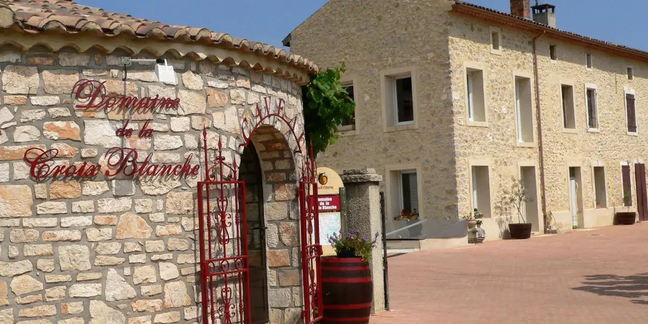 Bienvenue au **domaine de la Croix Blanche**. Une bâtisse en pierre avec une enseigne élégante et une porte ornée de ferronneries rouges.