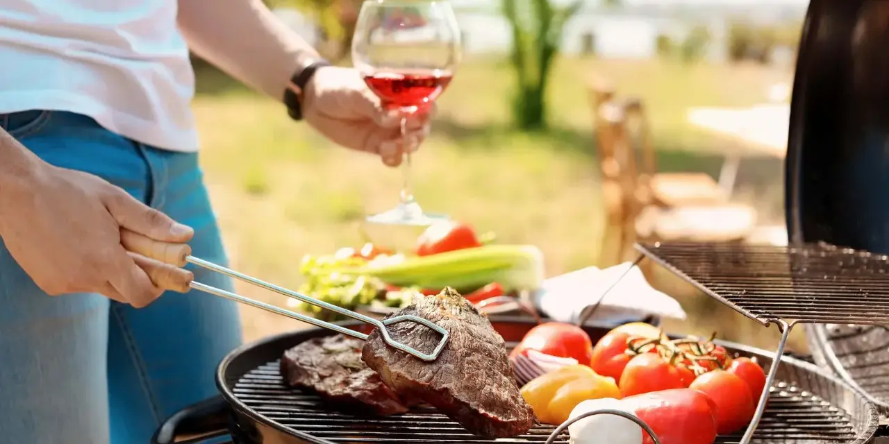 Un homme prépare un barbecue avec des steaks et des légumes. Il tient un verre de vin rouge, parfait pour accompagner ce repas. Quel vin avec barbecue ? Un rouge fruité !