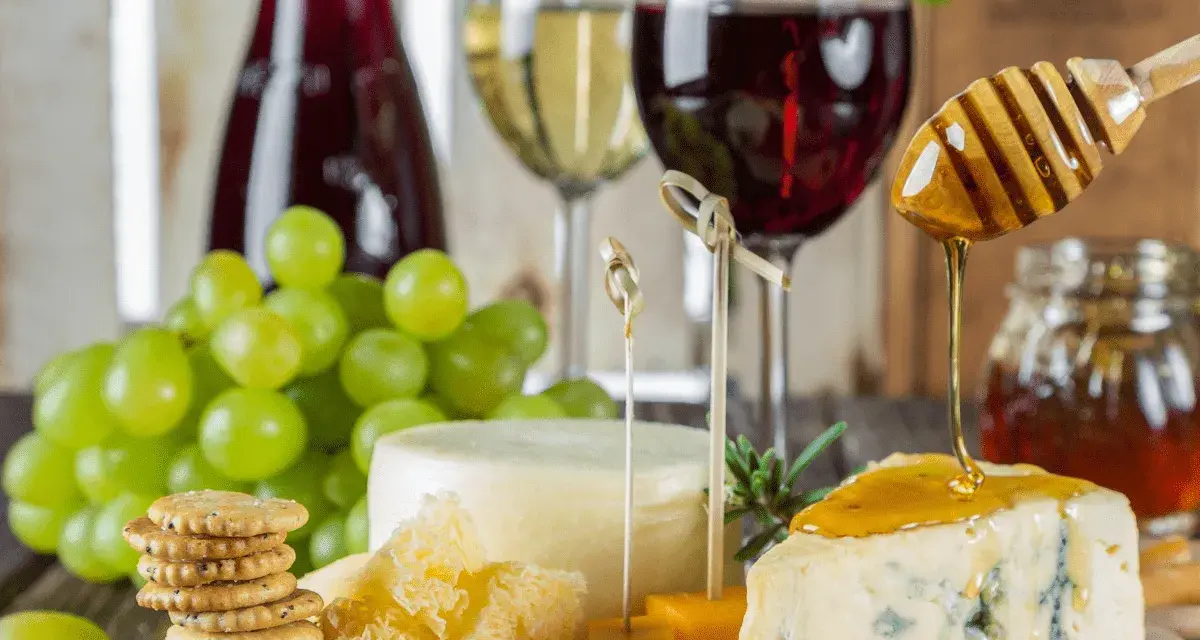 Un plateau de fromages, raisins verts, biscuits et miel dégoulinant sur un morceau de bleu, avec un verre de vin rouge léger en arrière-plan.