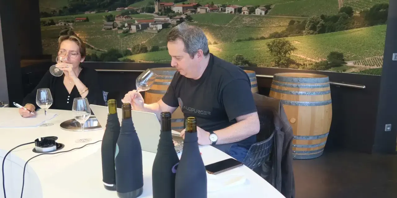 Dégustation de vins au domaine Mugneret Gibourg. Un homme et une femme goûtent du vin devant des bouteilles et un paysage de vignobles.