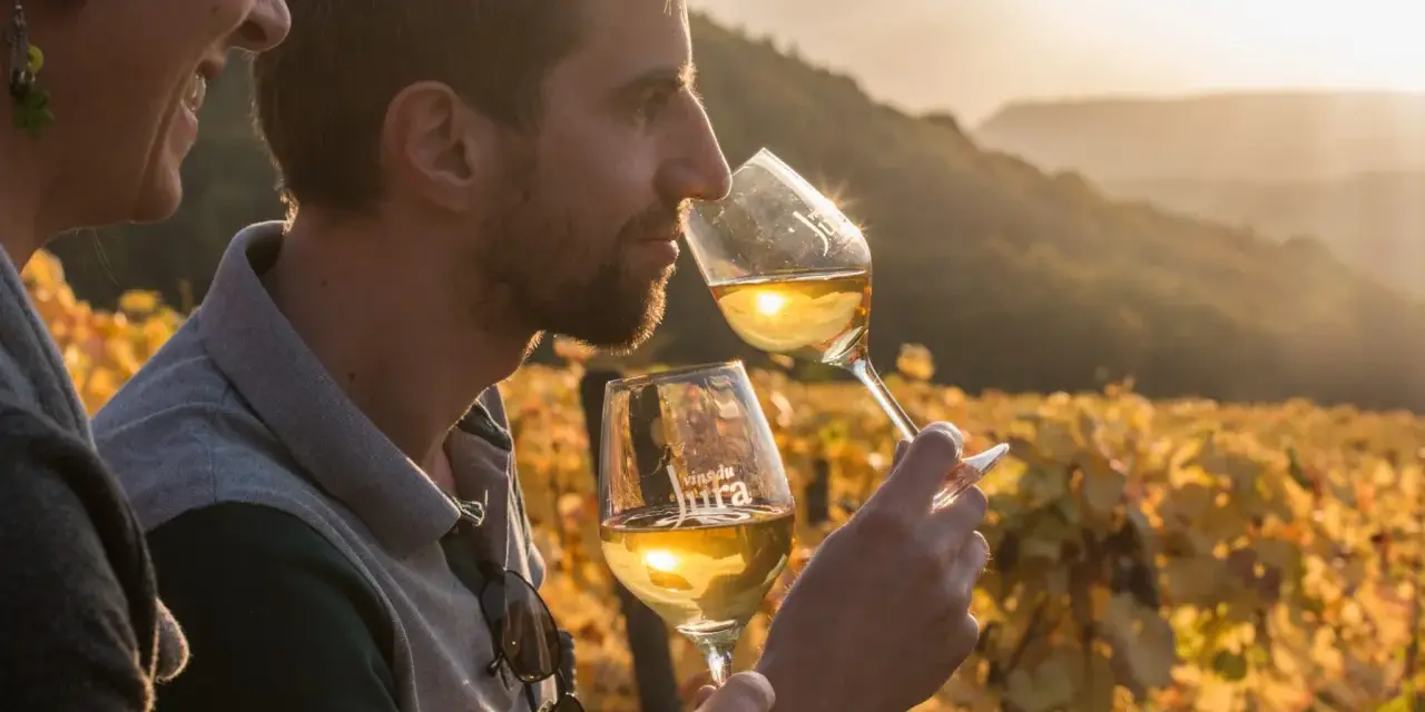 Un homme et une femme dégustent un vin jaune du Jura au coucher du soleil, dans un vignoble aux couleurs d'automne.
