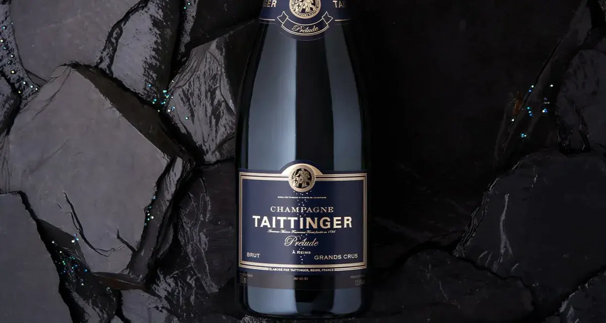 Bouteille de champagne Taittinger Prélude Grands Crus sur fond sombre, avec des paillettes scintillantes. Un avis sur ce champagne est à découvrir.