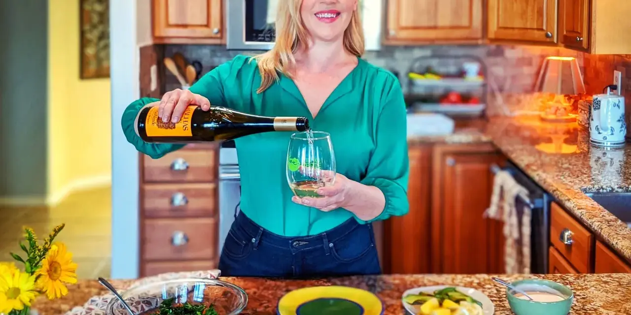 Une femme verse du vin vert dans un verre, avec une salade et des pains plats sur le comptoir.