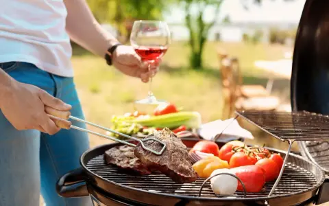 Un homme prépare un barbecue avec des steaks et des légumes. Il tient un verre de vin rouge, parfait pour accompagner ce repas. Quel vin avec barbecue ? Un rouge fruité !