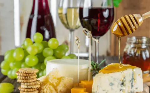 Un plateau de fromages, raisins verts, biscuits et miel dégoulinant sur un morceau de bleu, avec un verre de vin rouge léger en arrière-plan.