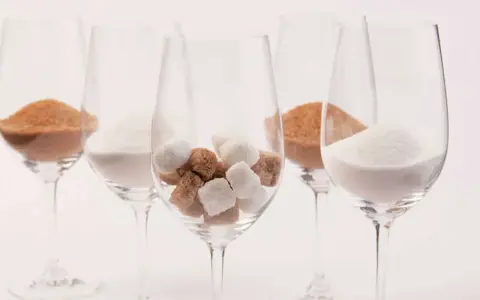 Cinq verres à vin contiennent différentes formes de sucre : du sucre en poudre blanc, du sucre brun en poudre et des morceaux de sucre blanc et brun. Une métaphore visuelle du sucre dans le vin.