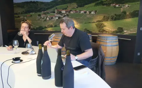 Dégustation de vins au domaine Mugneret Gibourg. Un homme et une femme goûtent du vin devant des bouteilles et un paysage de vignobles.