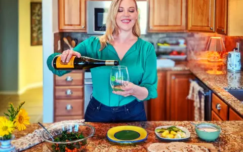 Une femme verse du vin vert dans un verre, avec une salade et des pains plats sur le comptoir.
