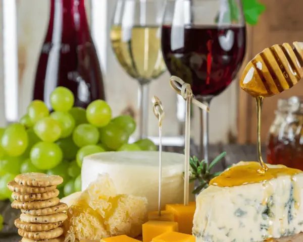 Un plateau de fromages, raisins verts, biscuits et miel dégoulinant sur un morceau de bleu, avec un verre de vin rouge léger en arrière-plan.