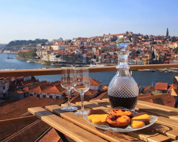 Verre de porto tawny ou ruby, deux verres à vin et des pâtisseries sur une table en bois, avec la ville de Porto en arrière-plan.