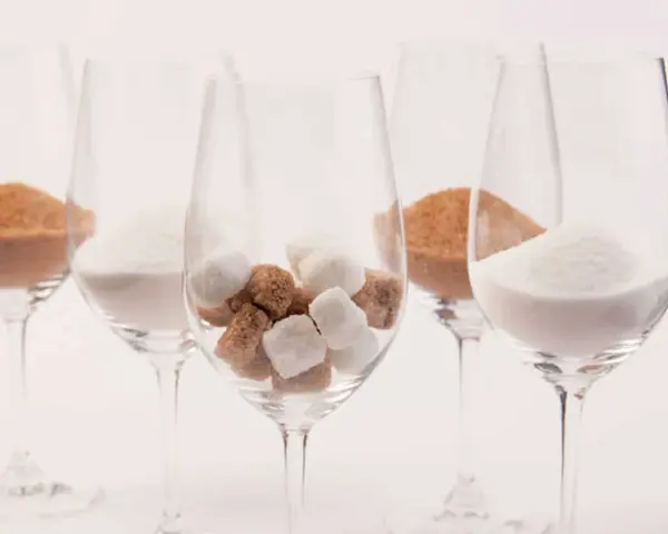 Cinq verres à vin contiennent différentes formes de sucre : du sucre en poudre blanc, du sucre brun en poudre et des morceaux de sucre blanc et brun. Une métaphore visuelle du sucre dans le vin.