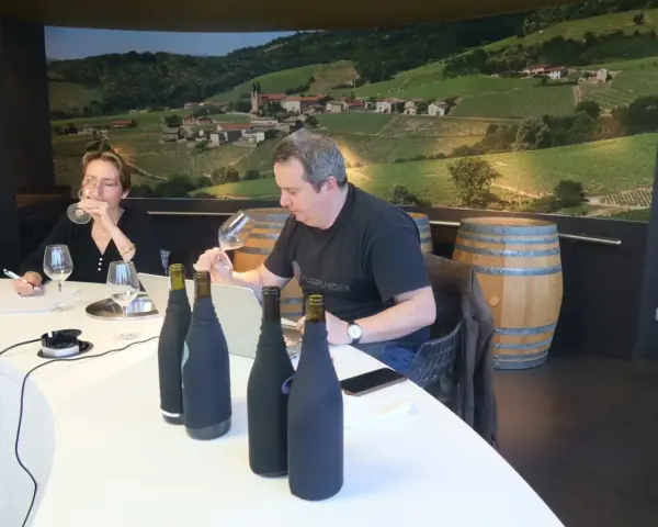 Dégustation de vins au domaine Mugneret Gibourg. Un homme et une femme goûtent du vin devant des bouteilles et un paysage de vignobles.
