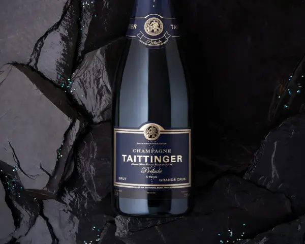 Bouteille de champagne Taittinger Prélude Grands Crus sur fond sombre, avec des paillettes scintillantes. Un avis sur ce champagne est à découvrir.