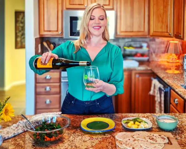 Une femme verse du vin vert dans un verre, avec une salade et des pains plats sur le comptoir.
