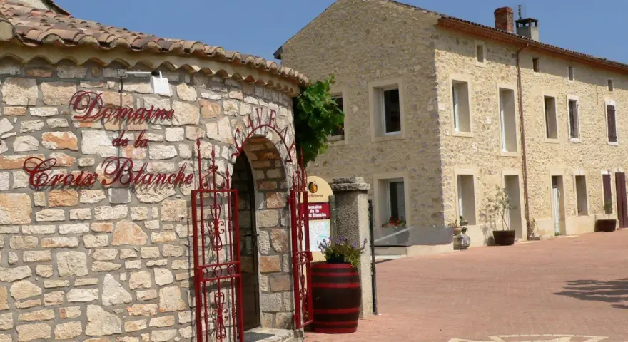 Bienvenue au **domaine de la Croix Blanche**. Une bâtisse en pierre avec une enseigne élégante et une porte ornée de ferronneries rouges.