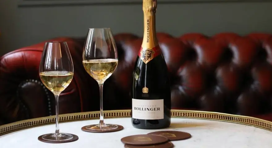 Bouteille de champagne Bollinger et deux verres remplis de champagne sur un plateau, avec un canapé en cuir rouge en arrière-plan.