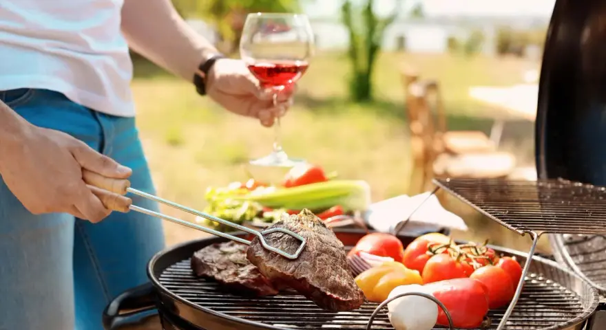 Un homme prépare un barbecue avec des steaks et des légumes. Il tient un verre de vin rouge, parfait pour accompagner ce repas. Quel vin avec barbecue ? Un rouge fruité !