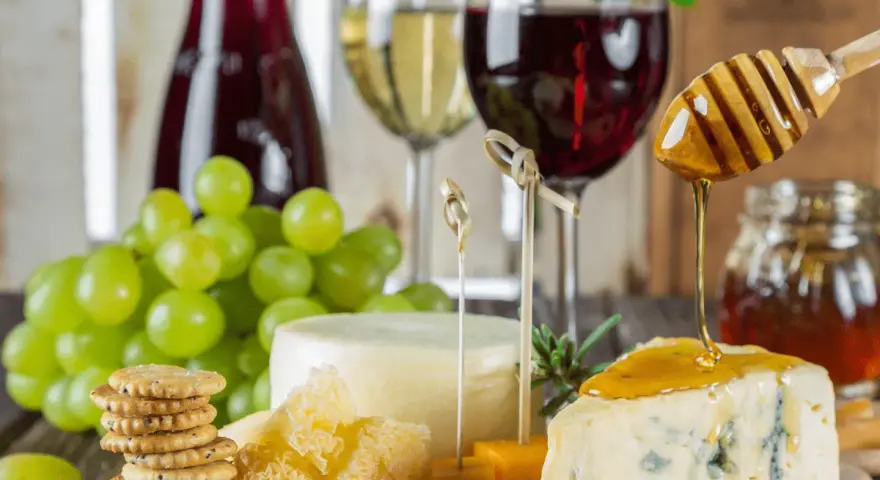 Un plateau de fromages, raisins verts, biscuits et miel dégoulinant sur un morceau de bleu, avec un verre de vin rouge léger en arrière-plan.