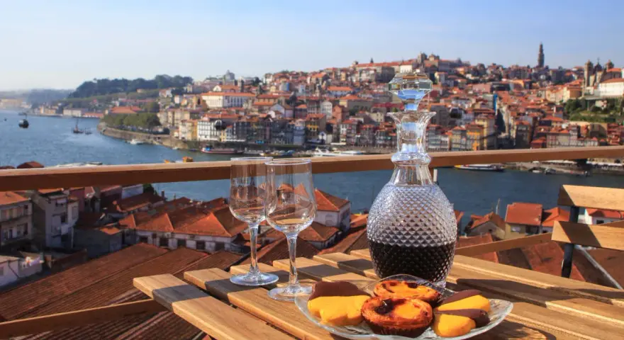 Verre de porto tawny ou ruby, deux verres à vin et des pâtisseries sur une table en bois, avec la ville de Porto en arrière-plan.