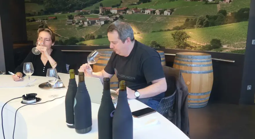 Dégustation de vins au domaine Mugneret Gibourg. Un homme et une femme goûtent du vin devant des bouteilles et un paysage de vignobles.