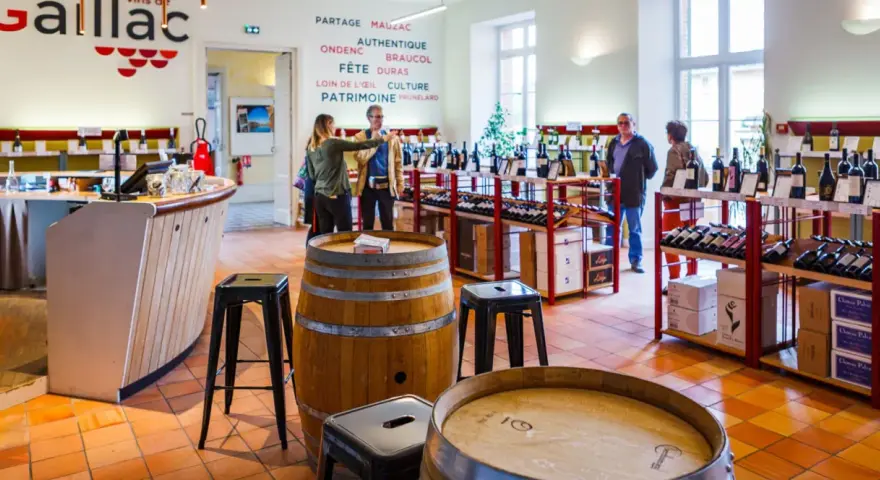Dans une cave à vin, des clients dégustent du vin du Tarn. Des étagères chargées de bouteilles et des tonneaux en bois complètent le décor.