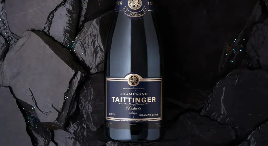 Bouteille de champagne Taittinger Prélude Grands Crus sur fond sombre, avec des paillettes scintillantes. Un avis sur ce champagne est à découvrir.