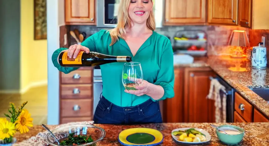 Une femme verse du vin vert dans un verre, avec une salade et des pains plats sur le comptoir.