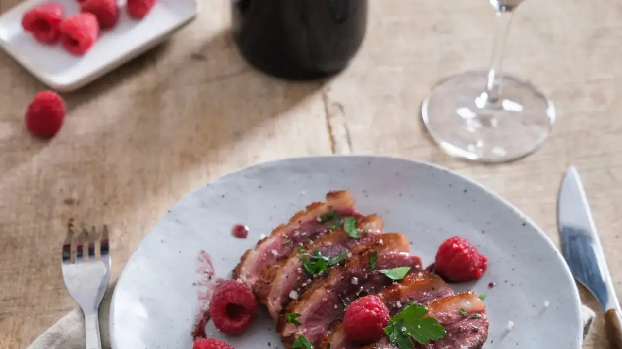 Tranches de magret de canard, garnies de framboises fra&icirc;ches et d'herbes, accompagn&eacute;es d'un verre de vin rouge.