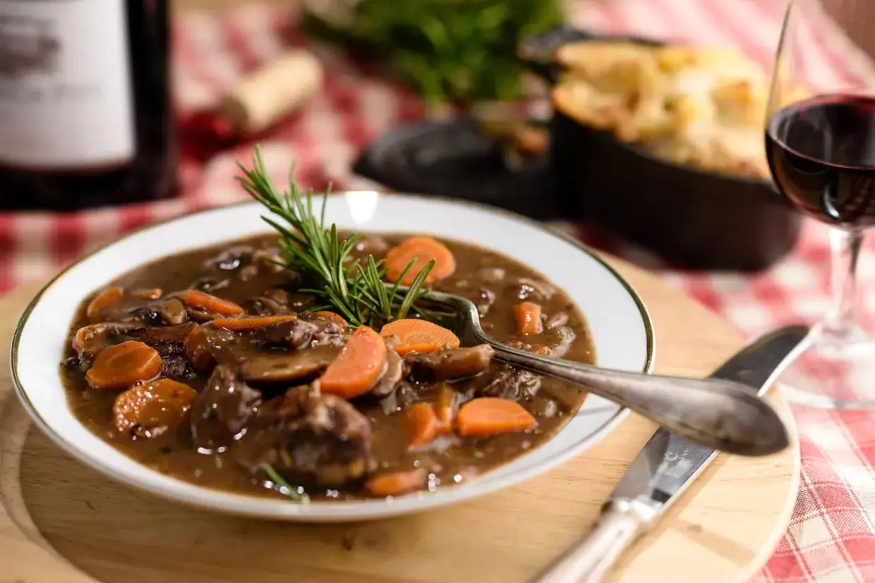 Un plat mijot&eacute; de b&oelig;uf aux carottes et champignons, garni de romarin, servi avec un verre de vin rouge.