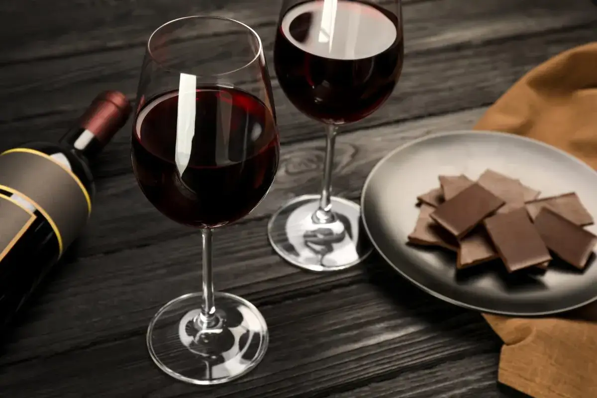 Deux verres de vin rouge, une bouteille et une assiette de chocolat sur une table en bois. Un accord parfait vin chocolat.