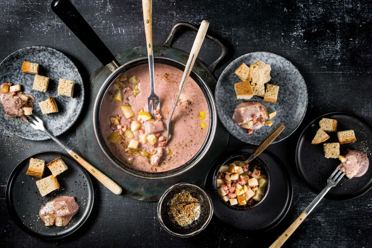 Fondue au vin rouge avec morceaux de poire, lardons et cro&ucirc;tons sur des assiettes noires et grises.